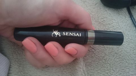 Eine Hand hält eine Tube Mascara von Sensai, die auf einem hellen Untergrund platziert ist.
