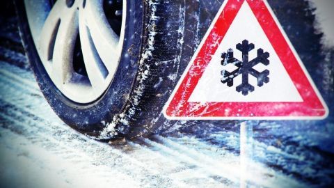 Ein Schneesymbol-Warnschild steht neben einem Autoreifen auf schneebedeckter Straße, geeignet für winterliche Bedingungen.