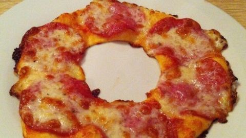Ein Pizza-Kranz mit geschmolzenem Käse und Tomatensauce liegt auf einem weißen Teller in einer hellen Umgebung.