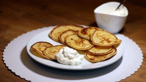 Auf einem weißen Teller liegen goldbraune Mini-Buttermilch-Pfannkuchen, daneben ein Schälchen mit Sahne, auf einem Tisch.