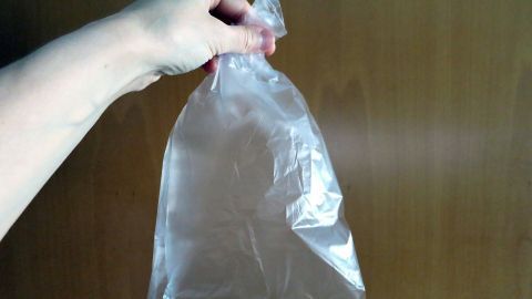 Eine Plastiktüte kann helfen, Schluckauf zu lindern. Atme in die Tüte ein und aus, um den CO₂-Gehalt zu erhöhen und so den Schluckreflex zu beruhigen. Achte darauf, dass die Tüte unbeschädigt ist und du sie richtig hältst. Eine Hand hält eine durchsichtige Plastiktüte vor einem holzfarbenen Hintergrund.
