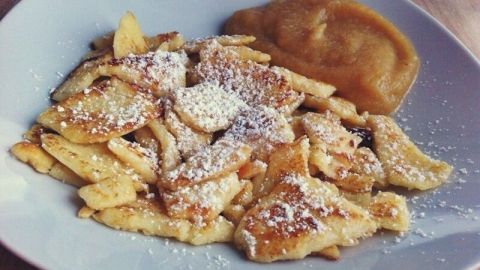 Fluffiger Kaiserschmarrn liegt auf einem weißen Teller, wird mit Puderzucker bestreut und hat eine Beilage aus Apfelmus.