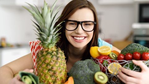 Eine lächelnde Frau umarmt frisches Obst und Gemüse, während sie in einer hellen Küche steht, was vegane Ernährung verdeutlicht.