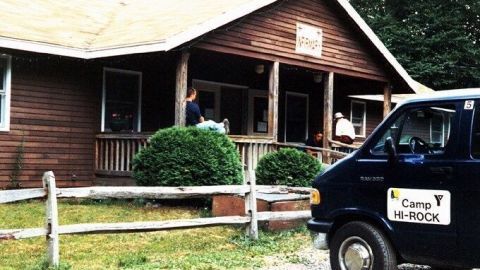 Ein Holzhaus steht vor einem grünen Baum, während ein Fahrzeug mit "Camp H-ROCK" parkt, ideal für Sommercamp-Aktivitäten.