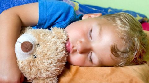 Ein schlafendes Kind hält einen Teddybären. Es liegt gemütlich im Bett mit bunter Bettwäsche. Das Bild vermittelt Ruhe.