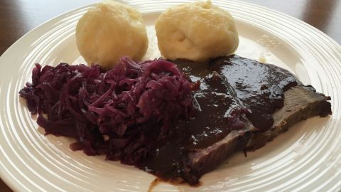 Kartoffelknödel liegen neben einem saftigen Stück Fleisch und Rotkohl auf einem weißen Teller in einer gemütlichen Umgebung.