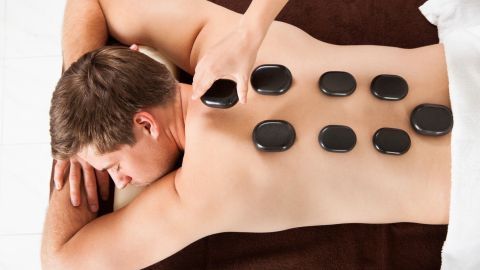Auf dem Rücken eines Mannes liegen glatte, warme Steine, während eine Hand einen weiteren Stein für eine Hot Stone Massage hebt.