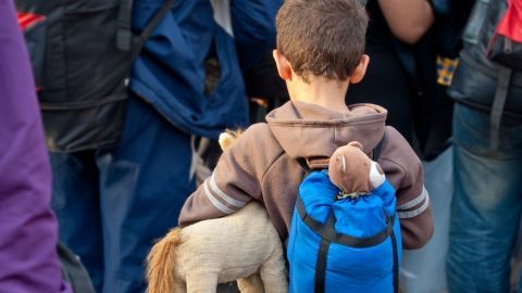 Ein kleiner Junge mit einem blauen Rucksack hält ein Stofftier und steht in einer Menschenmenge, die auf Hilfe wartet.