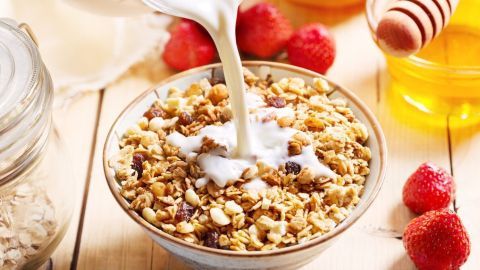 Eine Schüssel Müsli wird mit Milch gefüllt, umgeben von frischen Himbeeren und Honig im hellen Frühstücksbereich.