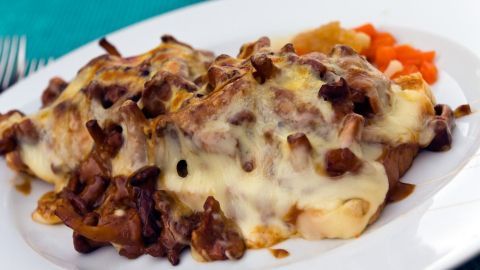 Ein überbackenes Schnitzel-Toast liegt auf einem weißen Teller, bedeckt mit geschmolzenem Käse, umgeben von Gemüse.