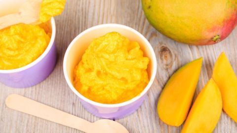 Leckeres Mangoeis wird in lila Schälchen serviert, umgeben von frischen Mango-Scheiben und einer ganzen Mango.