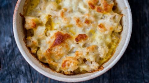 Ein Auflauf aus Blumenkohl, überbacken mit geschmolzenem Käse, steht in einer runden Schüssel auf einem dunklen Holzuntergrund.