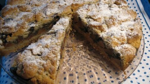 Eine Quitten-Mohn-Torte mit Butterstreuseln liegt auf einem Glasboden. Ein Stück wurde herausgeschnitten und die Torte ist mit Puderzucker bestäubt.