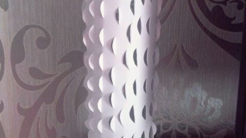 Eine weiße Vase mit ausgeschnittenem Muster steht auf einem Regal vor einer kunstvoll gemusterten Tapete.