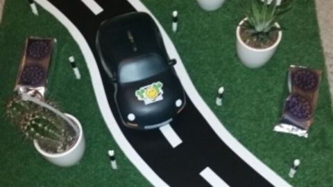 Ein schwarzes Spielzeugauto fährt auf einer grünen Fläche mit einer Straßen-Attrappe, umgeben von Töpfen und Süßigkeiten.