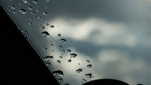 Wassertropfen liegen klar auf der Autoscheibe, während graue Wolken im Hintergrund sichtbar sind, die eine nasse Umgebung andeuten.