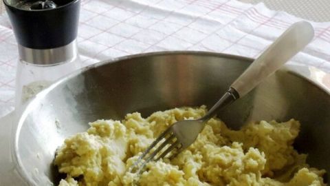 Ein metallischer Schüssel enthält Kartoffelpüree, während eine Gabel darin rührt; ein Salzstreuer steht daneben.