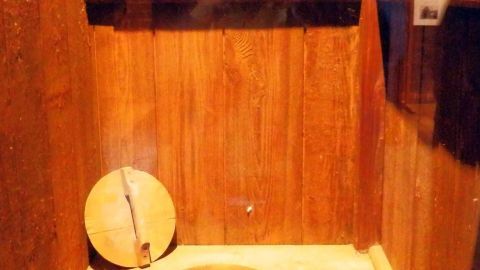 Eine einfache Holztoilette mit einem runden Loch, daneben eine Schale und darüber Regale mit grünen Pflanzen.