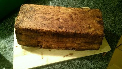 Ein Zimtkuchen mit Apfelstücken liegt auf einem Holzbrett, umgeben von einer dunklen, strukturierten Arbeitsfläche.