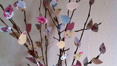 Zweige mit bunten Papierblättern zieren eine Vase, dekorativ arrangiert in einem hellen Raum.
