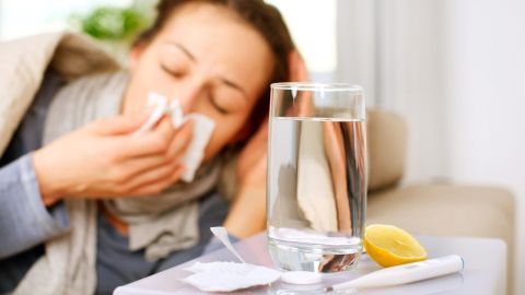 Eine kranke Frau sitzt im Wohnzimmer, putzt sich die Nase und trinkt Wasser gegen Husten und Schnupfen.