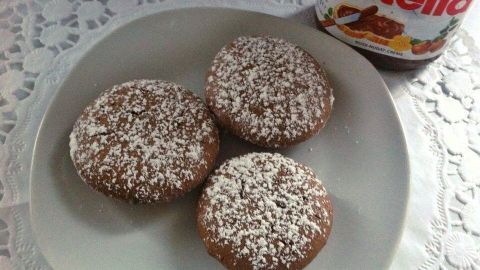 Auf einem Teller liegen drei mit Puderzucker bestäubte Nutella Küchlein. Daneben steht ein Nutella-Glas auf einem Spitzendeckchen.