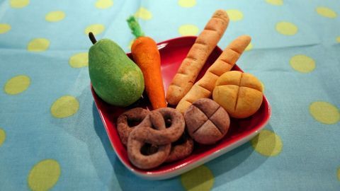 Bunte Salzteig-Objekte wie Gemüse und Brot in einer roten Schale auf einem hellen, gepunkteten Tisch.