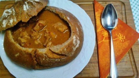 Ein Brotteiggefäß, gefüllt mit Gulaschsuppe, steht auf einem Holzbrett neben einem Löffel und einer serviettenartigen Dekoration.