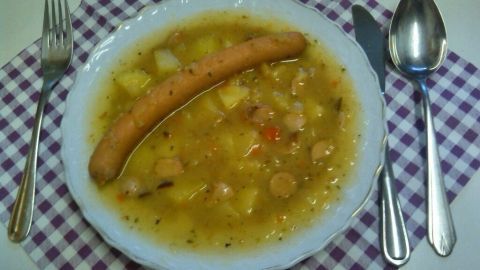 Eine heiße Schüssel Berliner Kartoffelsuppe mit Wurst liegt auf einem karierten Teller, umgeben von Besteck auf einem Tisch.