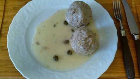 Zwei saftige Königsberger Klopse liegen in einer cremigen Sauce mit Kapern auf einem weißen Teller, umgeben von Holz.