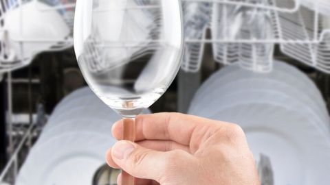 Ein Hand hebt ein klares Trinkglas hervor, während ein Geschirrspüler im Hintergrund Geschirr reinigt.