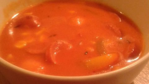Eine weiße Schüssel zeigt Cabanossi-Suppe mit oranger Brühe und Gemüse, steht auf einem Holztisch.