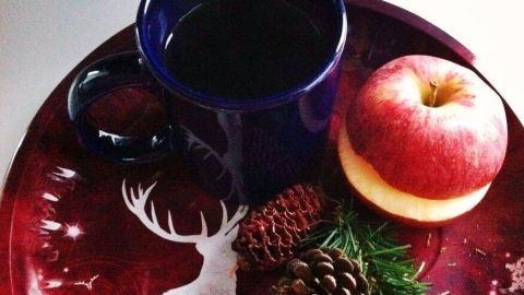 Ein blauer Becher mit Apfelpunsch steht auf einem festlichen Teller, umgeben von einem Apfel, Tannenzweigen und Tannenzapfen.