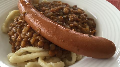 Eine Wurst liegt auf einem Bett aus Linsen und Spätzlen, serviert auf einem weißen Teller in einem gemütlichen Ambiente.