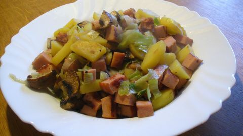 Leberkäse-Pfanne mit Lauch und Champignons auf einem weißen Teller, serviert auf einem Holztisch.