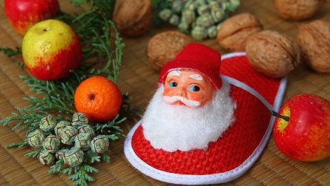 Nikolausfigur auf rotem Schuh, umgeben von Äpfeln, Mandarine, Nüssen und Tannenzweigen auf geflochtenem Untergrund.