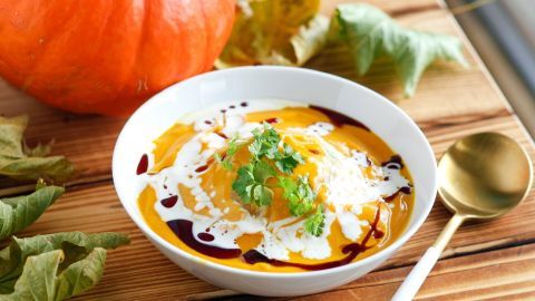 Ein weißer Teller mit cremiger Ingwer-Kürbissuppe, garniert mit frischem Koriander, steht neben einem Kürbis auf Holz.