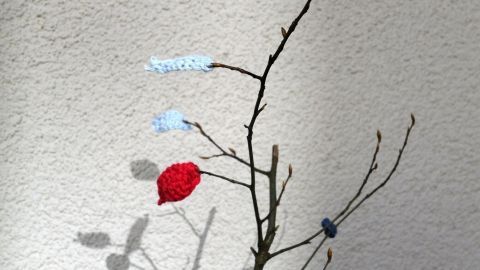 Bunte, gehäkelte Blätter schmücken einen dünnen Ast, der an einer hellen Wand steht und eine kreative Herbstdekoration bildet.