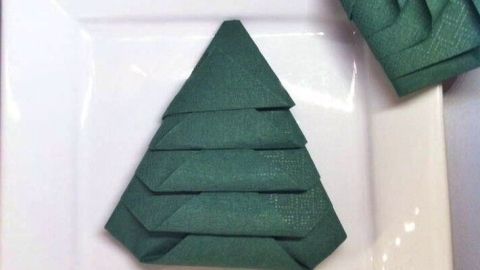 Ein gefalteter Servietten-Weihnachtsbaum ist auf einem weißen Teller platziert, neben einem weiteren Baum.