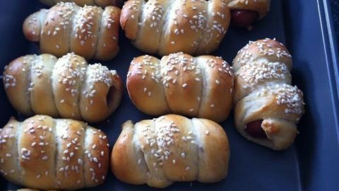 Goldbraune Hotdogs im Schlafrock liegen auf einem Backblech, gleichmäßig gebacken und mit Sesam bestreut.