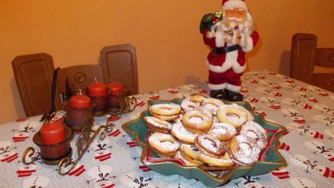Auf einem festlich gedeckten Tisch stehen auf einem Teller mit Marmelade gefüllte Plätzchen, umgeben von weihnachtlicher Dekoration.