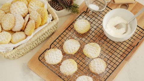 Auf einem Holzbrett liegen Puddingplätzchen, während Puderzucker durch ein Sieb darüber gestreut wird.