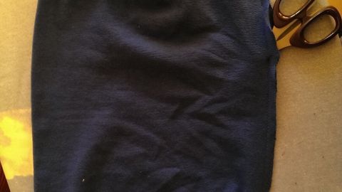 Dunkelblauer Stoffbeanie liegt auf Tisch, daneben eine Stoffschere. Hintergrund zeigt Holzboden und grünen Teppich.