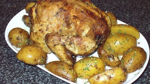 Ein goldenes Hähnchen liegt auf einem weißen Teller, umgeben von gebräunten Kartoffeln, auf einer dunklen Küchenoberfläche.