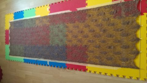 Ein Lacetuch wird auf bunten Schaumstoffplatten gespannt; dabei zeigt sich ein detailliertes Muster in einem Raum.
