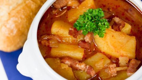 Ein weißer Topf mit Kartoffelgulasch, gewürzt mit Petersilie, neben einem frischen Brötchen auf einem blauen Untergrund.