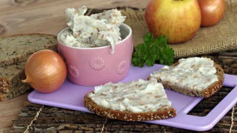 Eine Schale mit selbst gemachtem Apfel-Griebenschmalz steht neben belegten Broten, frischen Äpfeln und Zwiebeln auf einem Holzuntergrund.