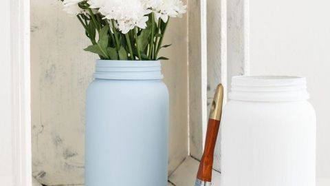 Ein graublauer Glasbehälter mit weißen Blumen steht in einer Holzkiste, während ein leerer weißer Behälter daneben liegt.