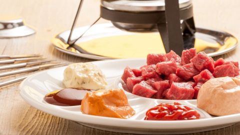 Ein Teller mit verschiedenen Fleischstücken und Saucen steht vor einem Fonduetopf, der auf einem Tisch platziert ist.