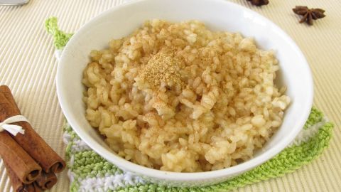 Eine Schüssel Milchreis ist mit Zimt bestreut, umgeben von Zimtstangen und Sternanissamen, auf einem Tischliegend.